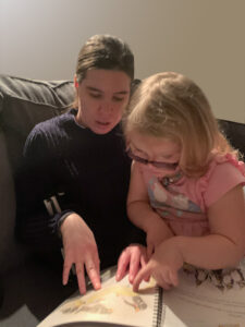 A blind woman in a blue sweater sitting on a couch with a young girl in a pink dress on her lap. They are sharing a print/Braille book.