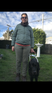 Shauna and her guide dog, Barney, with green grass and trees in the background. Shauna is wearing rose gold coloured sunglasses that have a clear amber tint to the lenses, khaki green cargo pants, brown leather ankle boots, and a hooded green puffer winter jacket with a dark grey turtleneck showing underneath. Barney, a male black Labrador retriever, is wearing a black gentle leader, Guide Dogs for the Blind harness, and a black collar with a brown leather leash.