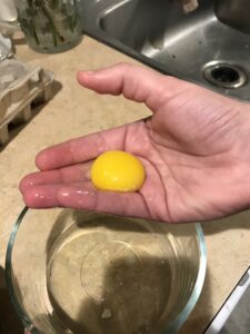 A hand, with an egg yolk in the palm. The fingers are slightly spread apart, so that the egg whites are dripping between the fingers into a bowl. In the background there is a kitchen counter, an egg carton, and a bowl containing egg whites.