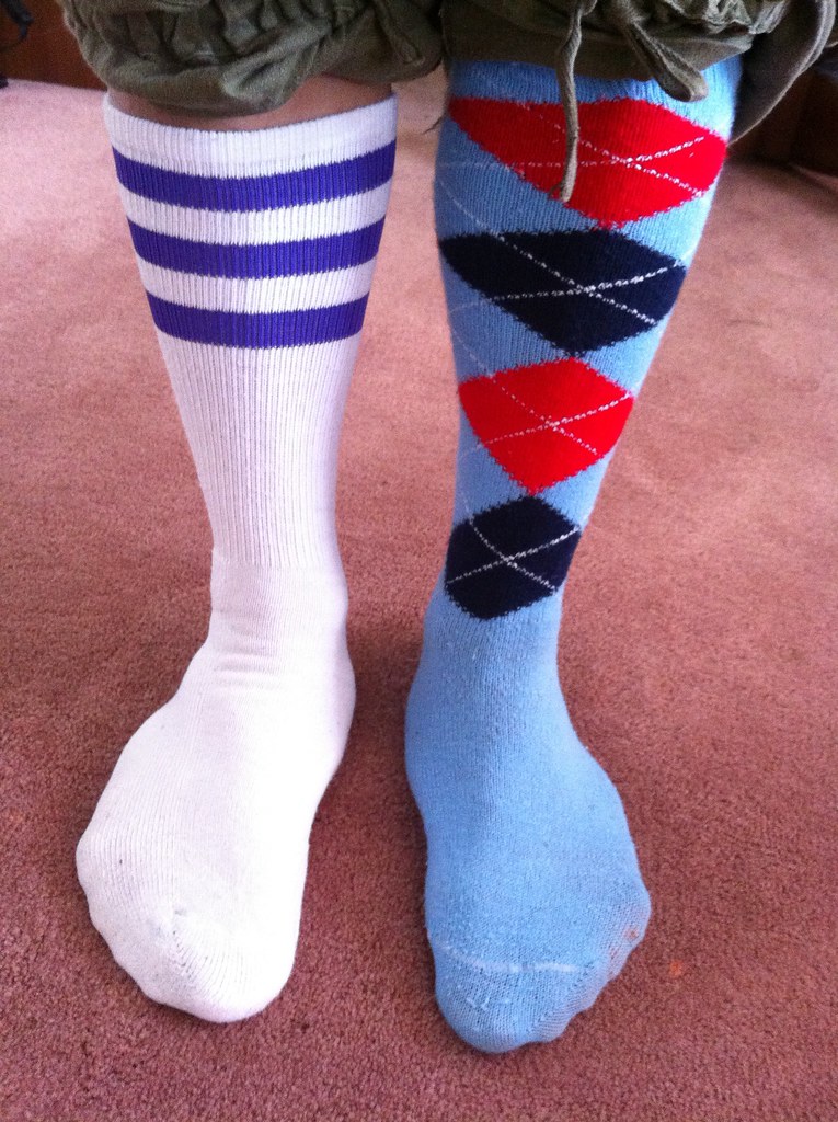 A pair of feet with mismatched socks. The sock on the left of the image is white with three dark blue stripes near the top. The sock on the right of the image is light blue and features four stacked diamonds. From top to bottom, the diamonds are red, dark blue, red, and dark blue.
